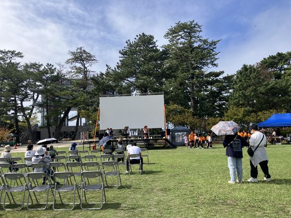 青空の下、松原公園の芝生に椅子を並べて開場を待つ会場（午前）