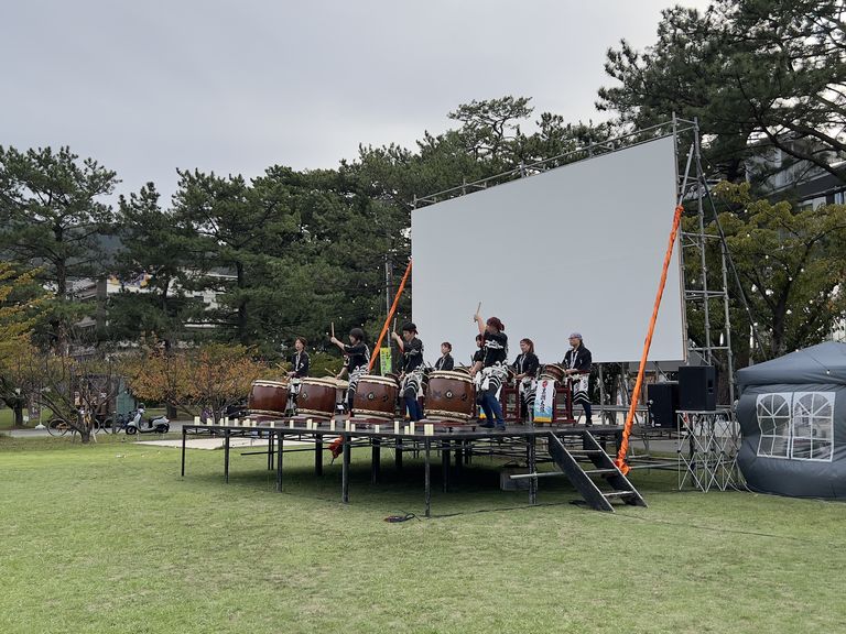 午後のステージでスクリーンを背に太鼓を打つ出演者たち