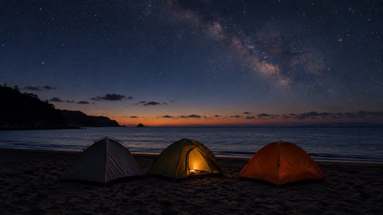 夜の砂浜に張られた3張りのドームテント。1張りの中にランタンの灯りがともり、上空に天の川がかかっている