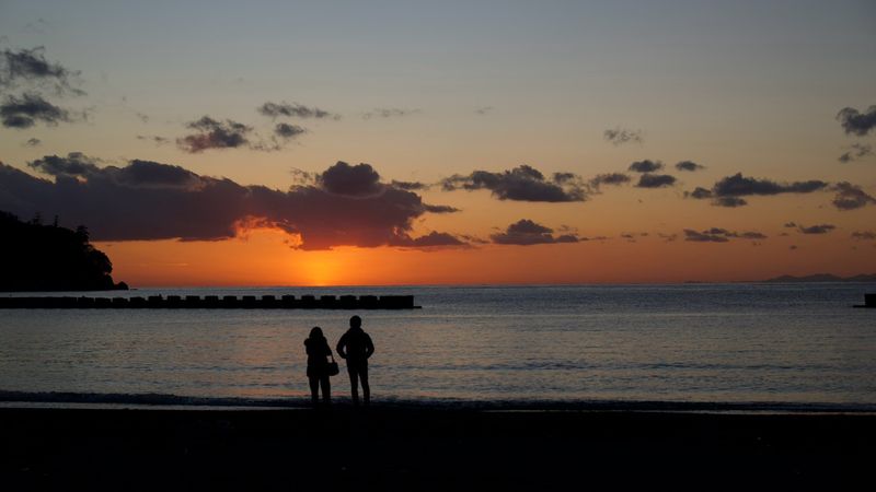 土肥の浜から見た夕暮れ。海に沈もうとする陽を、波打ち際に立つ二人が眺めている