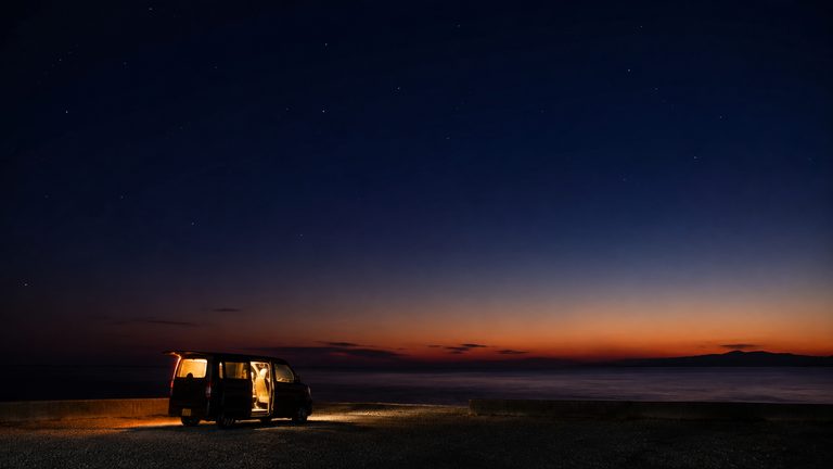 夜の海沿いの駐車場に一台だけ停まった車。ドアからもれる灯りが地面を照らし、上空には星が広がる