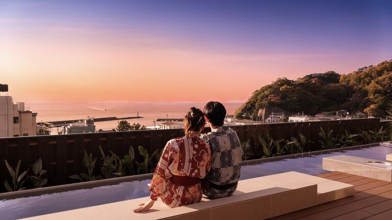 屋上の足湯テラス。浴衣姿の二人が腰かけて、夕焼けに染まる西伊豆の海と対岸の岬を眺めている