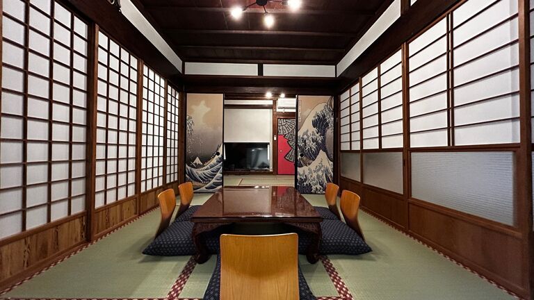 一棟貸しの和室の広間。障子に囲まれた畳の間に座卓が一列に並び、奥に海の絵が掛かっている