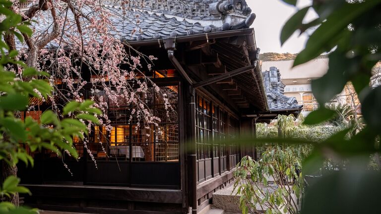 桜の枝ごしに見た、灯りのともる木造の宿の外観。瓦屋根と格子の縁側が緑に囲まれている