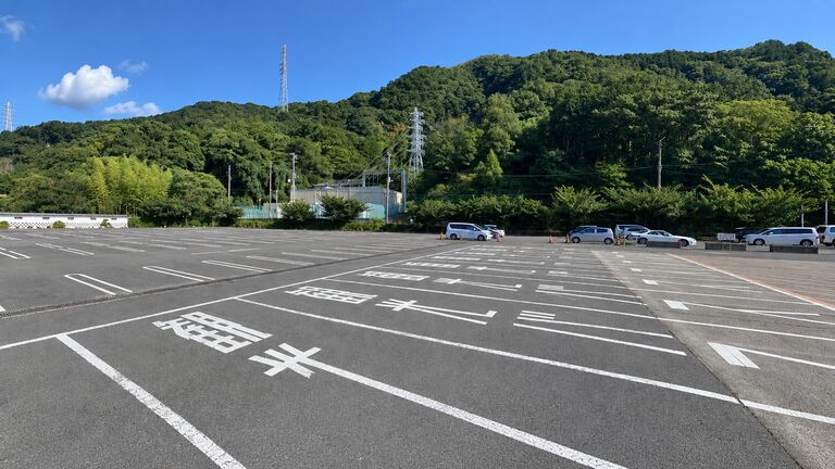 土肥金山の駐車場。白線の引かれた広い舗装面の奥に低い建物が建ち、その向こうに山並みと青空が見える