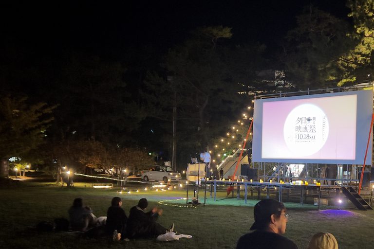 夜の松原公園で、電飾に囲まれて点灯した屋外スクリーンと、芝生に座って見つめる来場者たち
