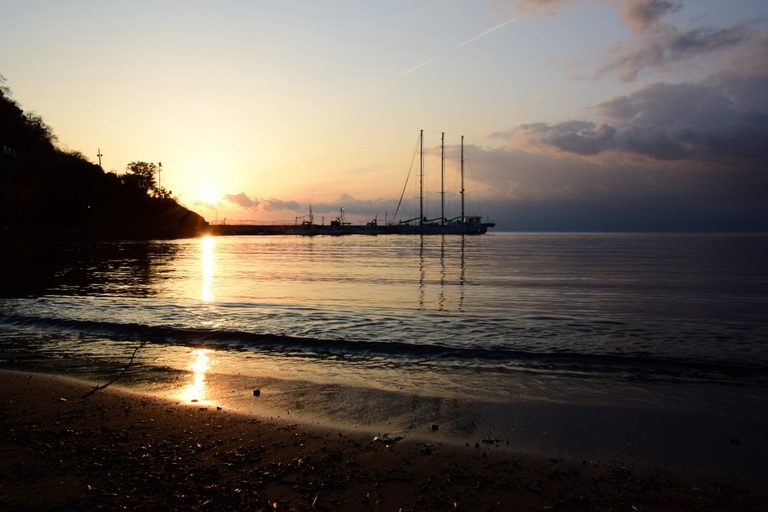 西伊豆・土肥の浜辺の夕暮れ。波打ち際に夕日が映り、沖に帆船が浮かんでいる