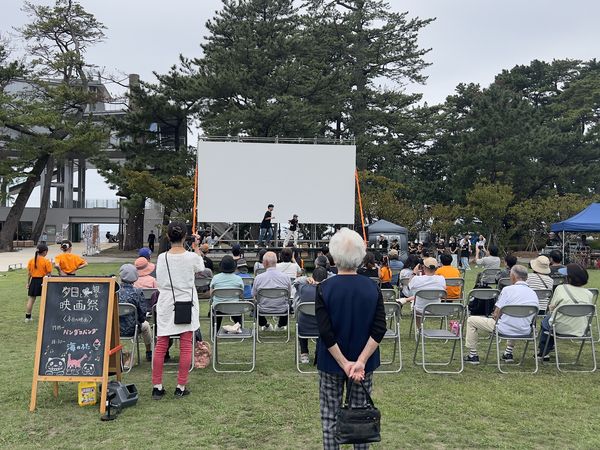 日中の松原公園で、椅子に座りマルシェのテントと屋外スクリーンを囲む来場者たち