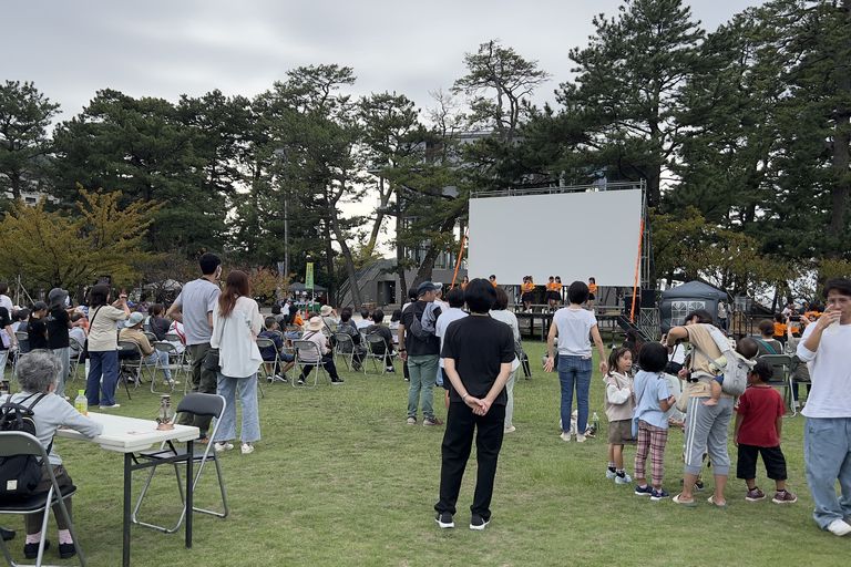 日中の松原公園で、出店のあいだを歩いたり腰かけたりする来場者と家族連れ