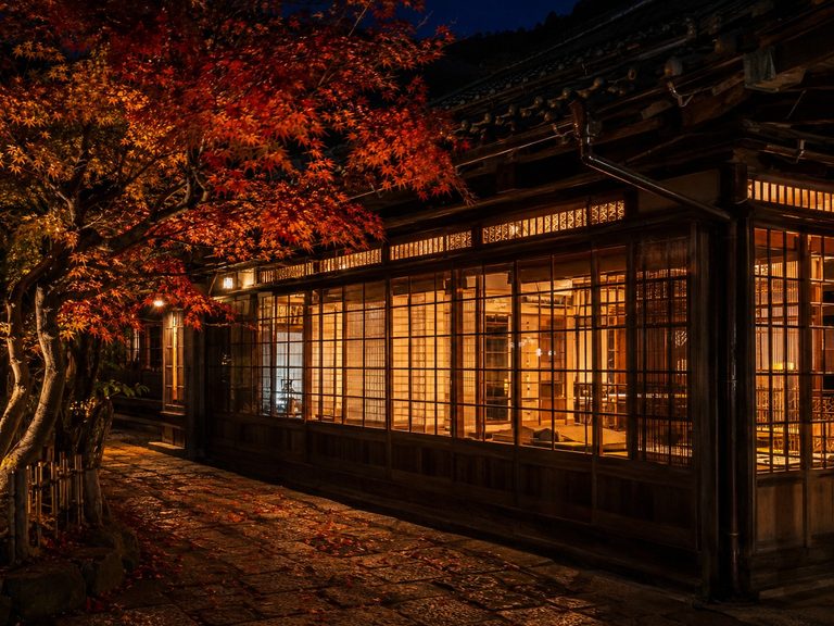 秋の夜の宿の外観。格子の引き戸一面から室内の暖かい灯りがもれ、手前に紅葉した木が枝を伸ばし、石畳に落ち葉がある