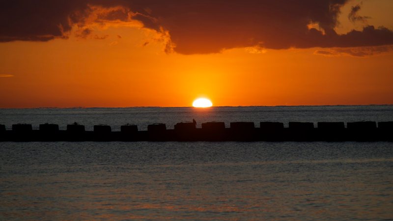 土肥の海に沈む夕日。オレンジの空の下、防波堤のシルエットの向こうで水平線に日が沈みかけている
